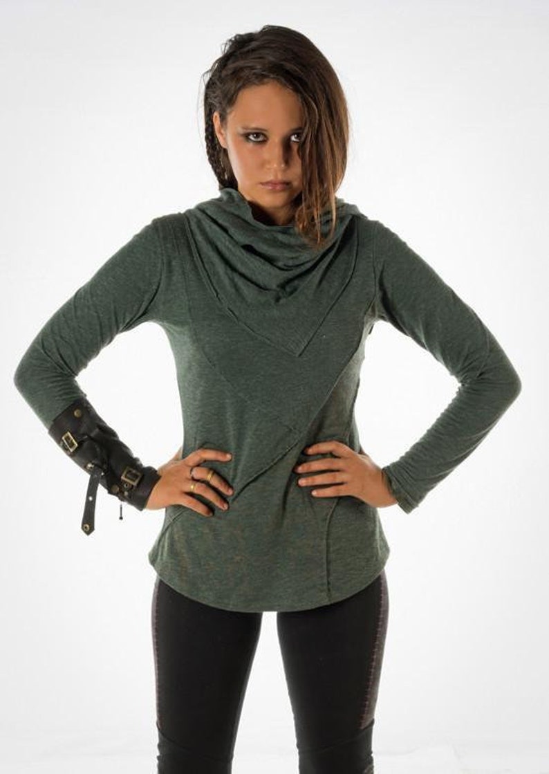 May include: A woman wearing a green long-sleeved shirt with a cowl neck and black pants. She is wearing a black leather cuff with metal accents on her left wrist.