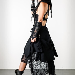 May include: A woman wearing a black hooded dress with lace detailing and black lace gloves. The dress has a layered skirt and a fitted bodice. She is wearing black boots.