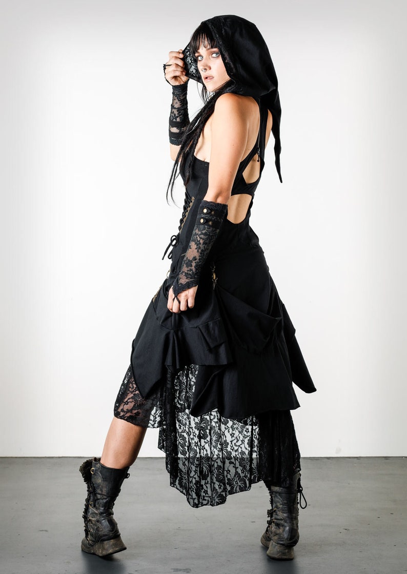 May include: A woman wearing a black hooded dress with lace detailing and black lace gloves. The dress has a layered skirt and a fitted bodice. She is wearing black boots.