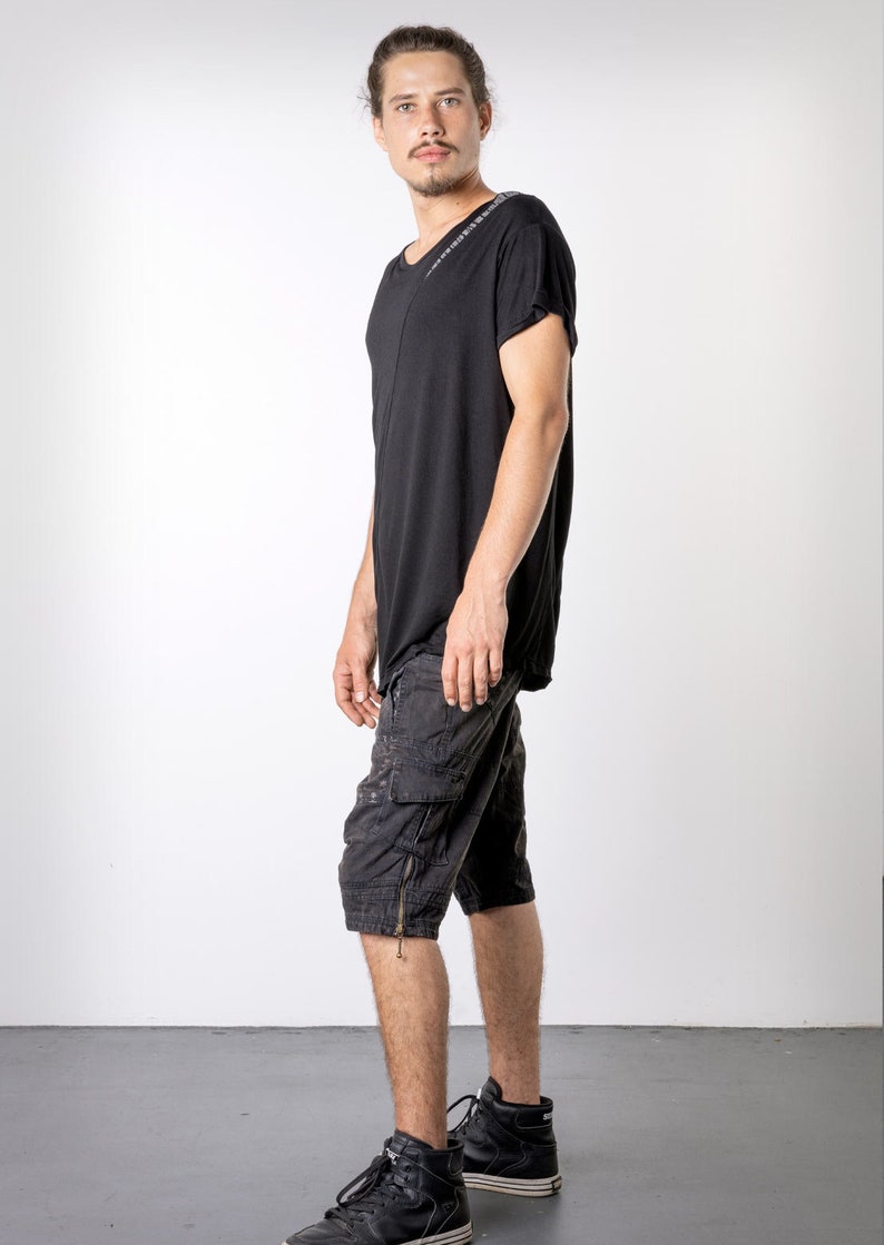 May include: Black t-shirt with a V-neck and short sleeves. The shirt is paired with black cargo shorts featuring multiple pockets and a zipper detail. The outfit is completed with black high-top sneakers. The model is standing in front of a white background.