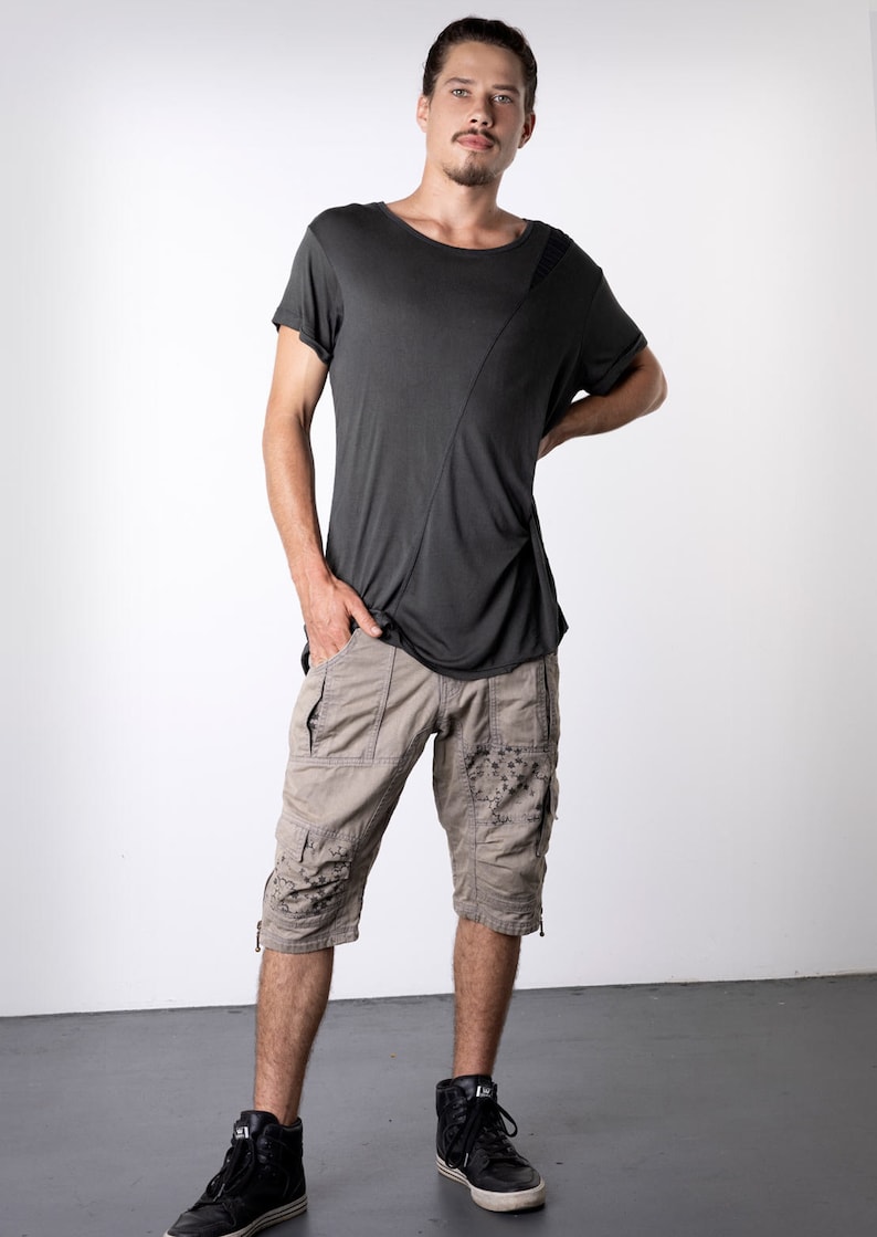May include: A dark gray short-sleeved t-shirt with a diagonal seam detail, paired with khaki cargo shorts featuring decorative embellishments. The outfit is completed with black high-top sneakers.