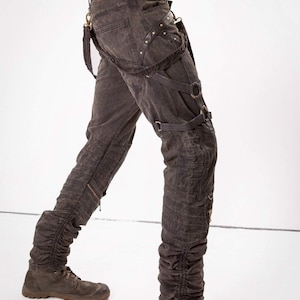 May include: Dark gray denim pants with multiple straps and buckles. The pants have a distressed look and are cuffed at the ankles. The model is wearing brown boots.