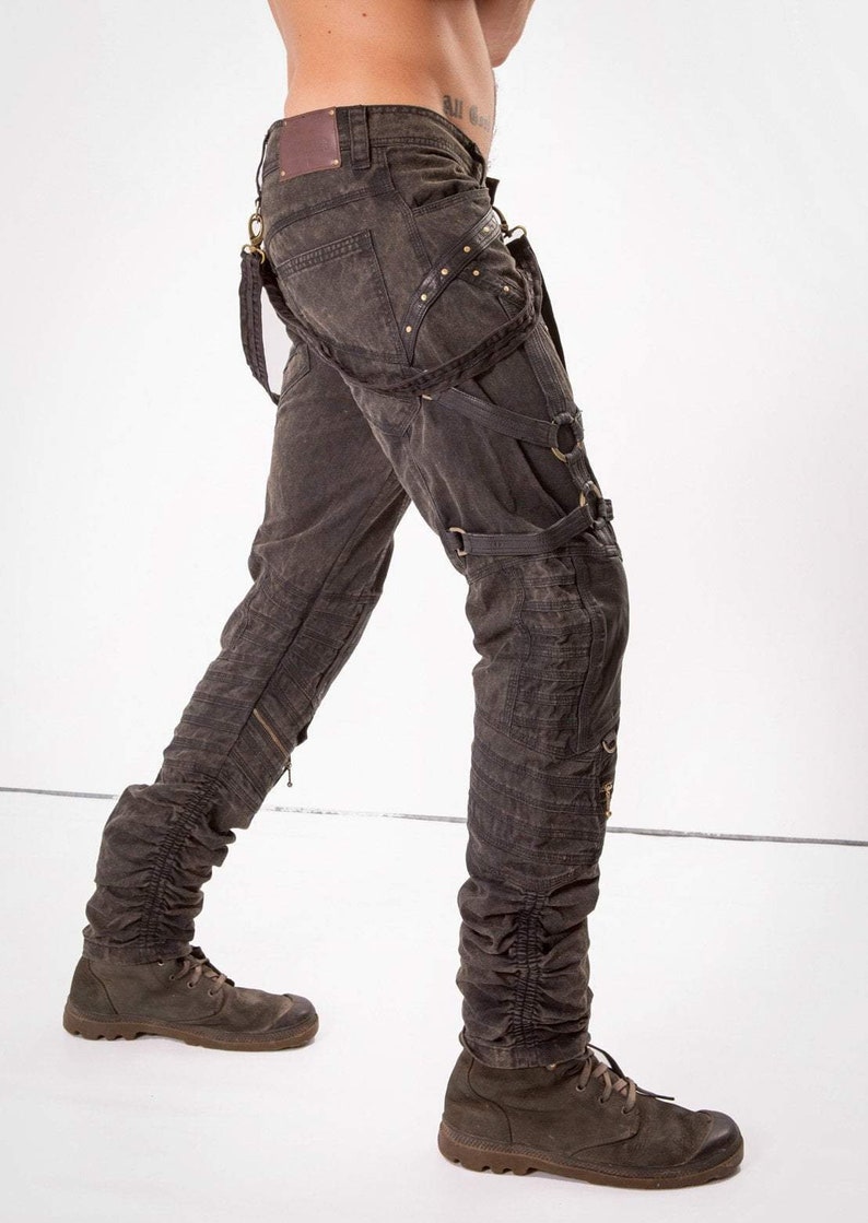 May include: Dark gray denim pants with multiple straps and buckles. The pants have a distressed look and are cuffed at the ankles. The model is wearing brown boots.
