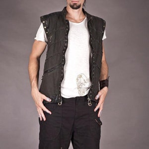 May include: A man wearing a black sleeveless vest with a white t-shirt underneath. The vest has a zip fastening and multiple pockets. He is also wearing black trousers with brown leather straps and goggles.