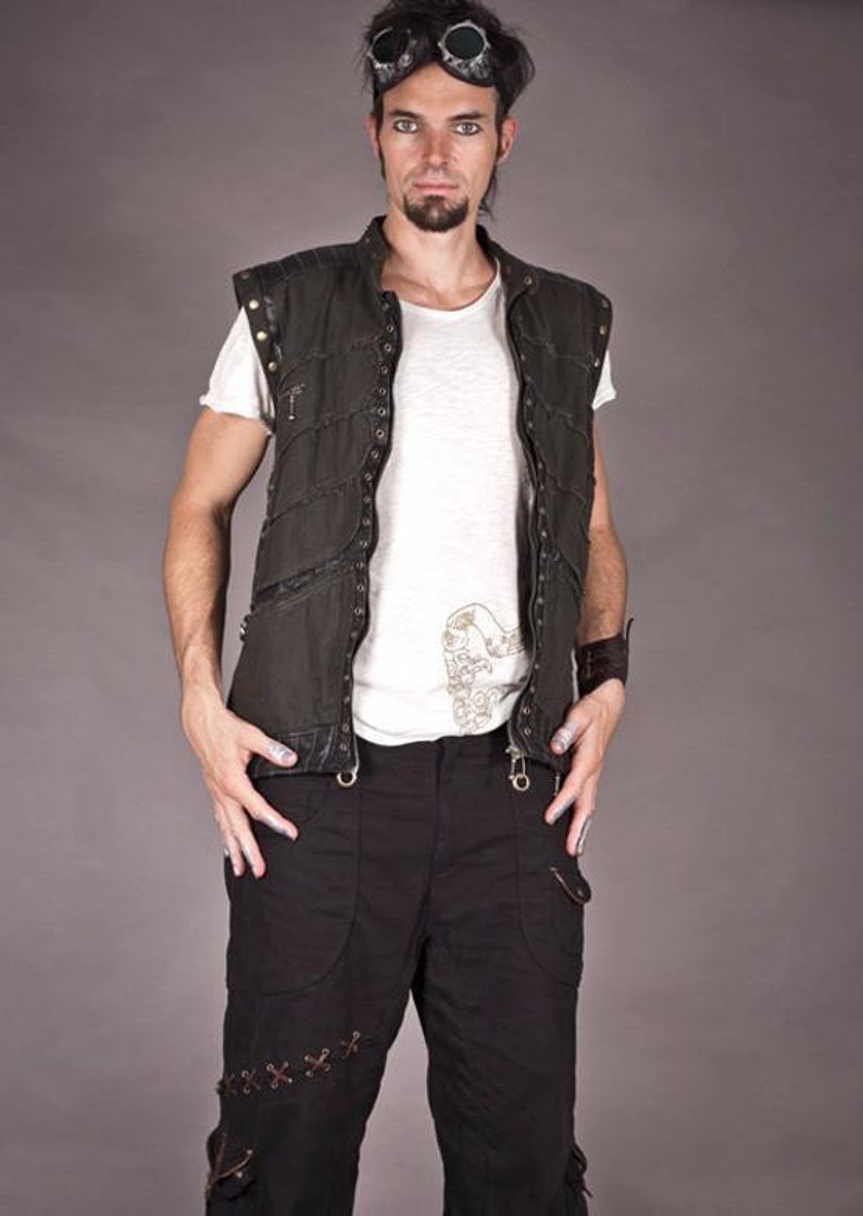 May include: A man wearing a black sleeveless vest with a white t-shirt underneath. The vest has a zip fastening and multiple pockets. He is also wearing black trousers with brown leather straps and goggles.