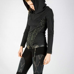 May include: A black long-sleeved hooded top with a green and black patterned design. The top has a high neckline and a fitted silhouette. The model is wearing black leggings with a geometric pattern.