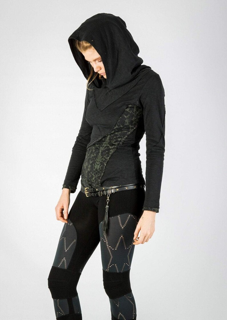 May include: A black long-sleeved hooded top with a green and black patterned design. The top has a high neckline and a fitted silhouette. The model is wearing black leggings with a geometric pattern.