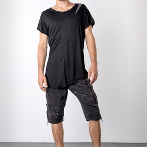 May include: Black asymmetrical t-shirt with a diagonal seam and a textured shoulder detail. The outfit includes black cargo shorts with multiple pockets and black high-top sneakers. The model is standing in front of a white background.