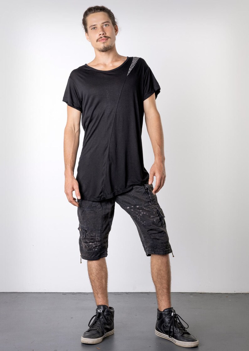 May include: Black asymmetrical t-shirt with a diagonal seam and a textured shoulder detail. The outfit includes black cargo shorts with multiple pockets and black high-top sneakers. The model is standing in front of a white background.