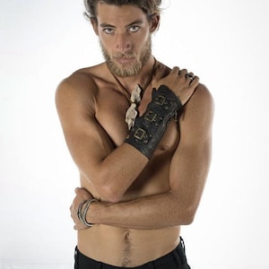 May include: A man with a beard and long hair is wearing a black leather wrist cuff with metal accents and a bone necklace. He is wearing black jeans and has his arms crossed.