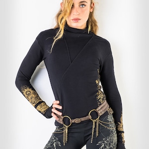 Könnte beinhalten: Ein schwarzes Langarmshirt mit goldenen Blumendesigns an den Ärmeln und einem braunen Ledergürtel mit goldenen Akzenten. Das Shirt hat einen Stehkragen und ein Wickeldesign. Das Model trägt eine schwarze Leggings mit goldenen Blumendesigns.