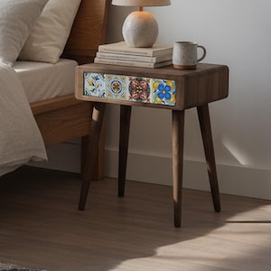 Op de afbeelding: Een houten nachtkastje met een lade met kleurrijke, patroon tegels. Bovenop staan een kleine lamp, boeken en een mok. De bedframe en het hoofdbord zijn zichtbaar op de achtergrond. De tafel is ongeveer 61 cm hoog.