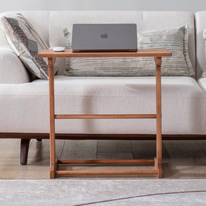 May include: A small, rectangular wooden side table with a laptop and mouse on top. The table is brown and has a simple, minimalist design. The table is placed in front of a white sofa with patterned pillows.