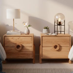 May include: Two wooden nightstands with a light finish, each featuring a drawer with a circular handle. One nightstand has a white lamp and books, the other a globe lamp. A bed with white bedding and a blue blanket is on each side.