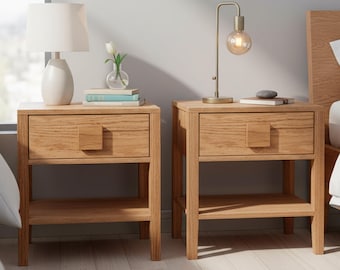 Può includere: Due comodini in legno chiaro, ciascuno con un cassetto e un ripiano inferiore. Uno ha una lampada bianca, l'altro una lampada dorata. I comodini sono posizionati accanto a letti con biancheria da letto bianca.