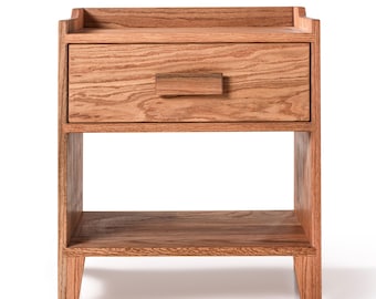 Puede incluir: Una mesita de noche de madera clara con un cajón y un estante abierto. La madera muestra vetas visibles y un tirador rectangular. La parte superior tiene un borde elevado.