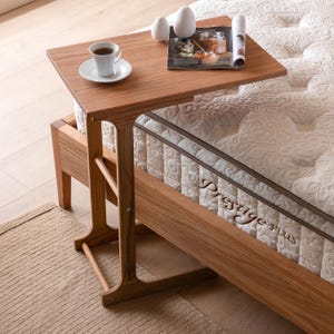 Op de afbeelding: Een lichtbruine houten bijzettafel met een rechthoekig blad, met een kopje koffie op een schotel, een tijdschrift en twee decoratieve eieren. De tafel staat naast een bed met een witte matras.