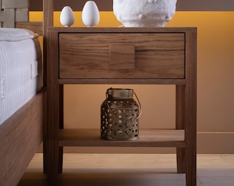 Puede incluir: Una mesita de noche de madera con un cajón y un estante inferior. En la parte superior, hay una lámpara blanca texturizada y dos decoraciones en forma de huevo. Una linterna decorativa de metal se encuentra en el estante inferior.