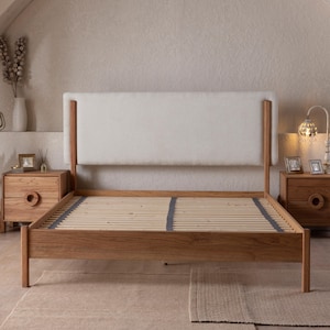 May include: A wooden bed frame with a light-colored upholstered headboard. The bed frame is made of light-colored wood and features a slatted base. Two wooden nightstands with circular drawer pulls flank the bed, each with a lamp and decorative items.