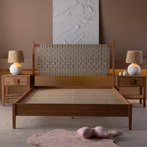 May include: A wooden bed frame with a woven headboard. Two matching wooden nightstands with lamps are on either side of the bed. The lamps have textured white bases and tan shades. A pink rug and pillows are on the floor.