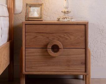 Può includere: Un comodino in legno a due cassetti con una maniglia circolare su ogni cassetto. Il comodino è realizzato in legno chiaro e ha una foto incorniciata sopra. Una lampada bianca con una base decorativa è sul comodino.