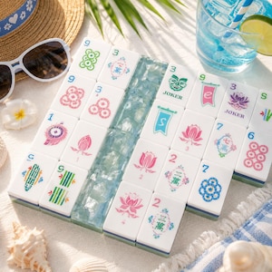 May include: A collection of white Mahjong tiles featuring colorful floral, numerical, and character designs, arranged in a grid pattern with a central column of translucent blue glass tiles, displayed alongside a straw hat and sunglasses.