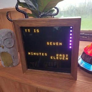 Puede incluir: Un reloj digital con marco de madera muestra la hora: "TT IS SEVEN MINUTES PAST ELEVEN" en letras amarillas iluminadas. Una estatua decorativa de búho y una lámpara roja también son visibles.
