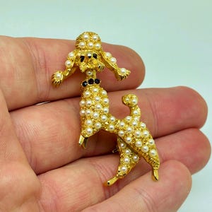 Pode incluir: Broche de poodle dourado adornado com pérolas falsas. O corpo e a cabeça do poodle são cobertos por pequenas pérolas redondas, com uma coleira preta. O broche é segurado em uma mão, mostrando seu design intrincado e estilo vintage.