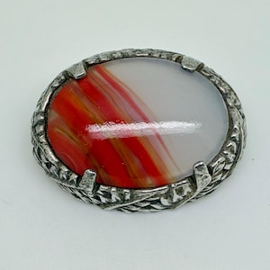 Könnte beinhalten: Ovale Vintage-Brosche mit silberfarbenem Metallrahmen. Die Brosche hat einen polierten Stein mit roten, orangefarbenen und weißen Wirbelmustern. Der Rahmen hat ein strukturiertes, blattartiges Design.