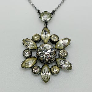Può includere: Una collana con una montatura in metallo scuro caratterizzata da un cristallo rotondo trasparente centrale circondato da cristalli trasparenti a forma di marquise e rotondi in un motivo a stella, con un ulteriore cristallo rotondo in cima.
