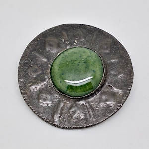May include: A round, antique silver-toned brooch with a central, large, green, polished stone. The brooch has a textured surface with etched designs around the stone. The stone is a vibrant, glossy green.