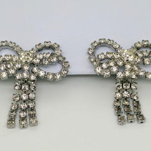 Puede incluir: Dos broches plateados con forma de lazo, incrustados con brillantes transparentes. Cada broche presenta un gran brillante central y hileras de brillantes colgantes, creando un accesorio brillante y elegante.