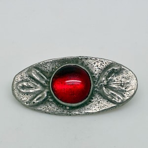 May include: Oval-shaped silver-toned brooch with a large, round, red gemstone center. The brooch has a textured surface and leaf-like designs on each end. A vintage-style accessory.