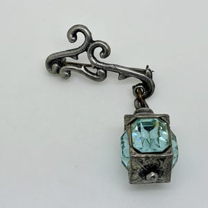 May include: A vintage silver-toned brooch featuring a decorative lantern design. The brooch has a stylized, ornate top with a dangling lantern pendant. The lantern is square-shaped and contains light blue, faceted crystals.