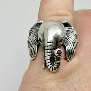 Peut inclure: Bague argentée en forme d'éléphant avec une trompe, des oreilles et une tête détaillées. La bague comporte une petite pierre précieuse rouge et ronde pour un œil. La bague est portée sur un doigt.