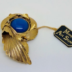 Puede incluir: Broche vintage dorado con una gran piedra azul ovalada en el centro. El broche tiene un diseño de hojas y una etiqueta que dice "Mony Art Style".