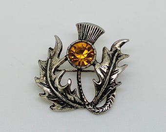 Puede incluir: Un broche de metal plateado con forma de flor de cardo. El centro del cardo presenta una piedra preciosa redonda y facetada de color ámbar. El metal tiene un acabado envejecido con patrones detallados de hojas espinosas alrededor.