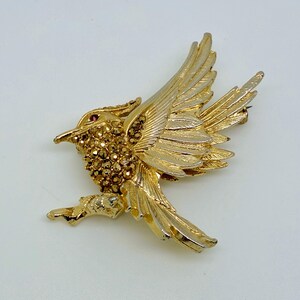 May include: A gold-coloured bird brooch in flight, with detailed feathering and a textured body. The bird's body is encrusted with small, sparkling amber-coloured stones, and it has a small red stone for an eye. The brooch is set against a white background.