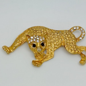 May include: Gold-coloured metal brooch depicting a crouching leopard. The brooch is covered in small, textured circles and features black eyes and clear crystal accents. A stylish accessory.