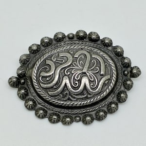 May include: Oval-shaped silver-toned metal brooch with an ornate, swirling design in the center. The brooch is surrounded by a rope-like border and small, rounded embellishments. The brooch is set against a white background.