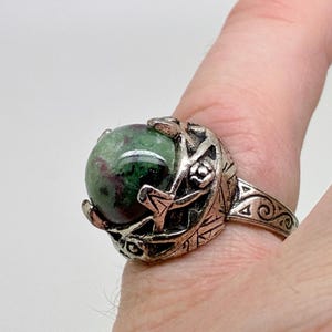 May include: A silver-toned ring featuring a large, round gemstone with green and red hues. The ring's setting has an ornate, textured design with floral and geometric elements. The band has a detailed, swirling pattern.
