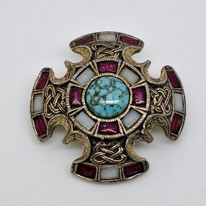 May include: A cross-shaped brooch with an antique silver-tone finish featuring a central round turquoise-colored stone with dark veining. The design includes inlaid rectangular magenta and white stones, accented by intricate raised knotwork patterns.