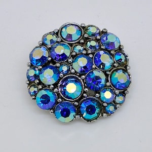 Puede incluir: Broche redondo con un grupo de brillantes de color azul y verde iridiscente. Los brillantes están engastados en un marco de metal plateado. El broche tiene un diseño clásico y elegante.