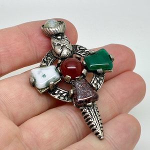 May include: A silver brooch with a decorative design, featuring a central red stone, a green stone, a white stone, and a brown stone. The brooch has an ornate, detailed pattern and a pin closure.
