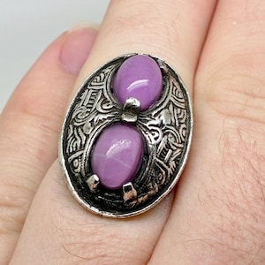 May include: Oval-shaped silver ring with two oval, light purple stones. The ring has an ornate, engraved design around the stones. The ring is on a finger.