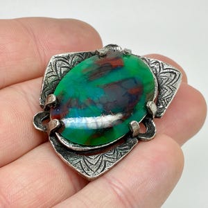 May include: A silver brooch featuring an oval, iridescent green and red stone. The stone is set in a decorative frame with a floral pattern. The brooch is held in a hand.