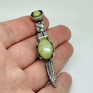 May include: A silver pendant featuring a light green oval gemstone. The pendant has an ornate design with a smaller green stone at the top and a textured, elongated base. The pendant is held in a hand against a white backdrop.