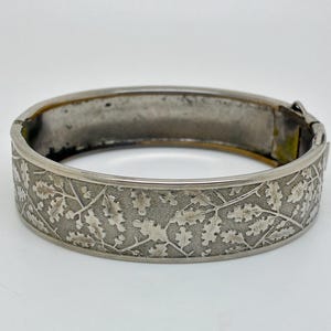 Puede incluir: Una pulsera rígida articulada de metal plateado con un patrón en relieve de hojas de roble y pequeñas bellotas en la superficie exterior. La pulsera tiene un acabado interior liso y un cierre mecánico seguro en el lateral.