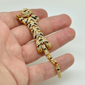 Peut inclure: Broche dorée en forme de tigre avec des rayures noires et des accents de cristaux transparents. La broche représente un tigre couché, la queue tendue. La broche est probablement en métal et est un accessoire décoratif.