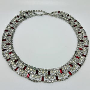 May include: A silver-tone metal collar necklace featuring a multi-layered design of clear round rhinestones and rectangular red baguette crystals. The piece is constructed with a flexible lattice framework and includes a metal hook clasp closure.
